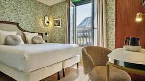 Habitación de hotel con cama, una silla, una mesa, un cuadro en la pared y ventana con vistas a un edificio.