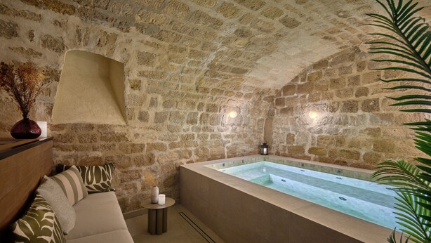 Una habitación con paredes de piedra, una bañera grande, un banco de madera y una mesita con una vela.