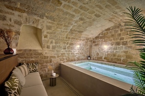 A stone-walled room with a large bathtub, a wooden bench, and a small table with a candle. - Hôtel Joséphine & SPA by HappyCulture (Paris)
