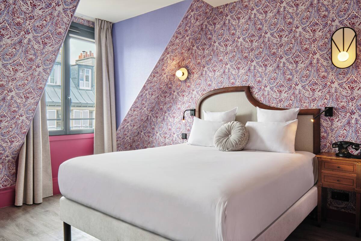 Un dormitorio con cama, mesita de noche, luz en la pared y ventana con cortinas.