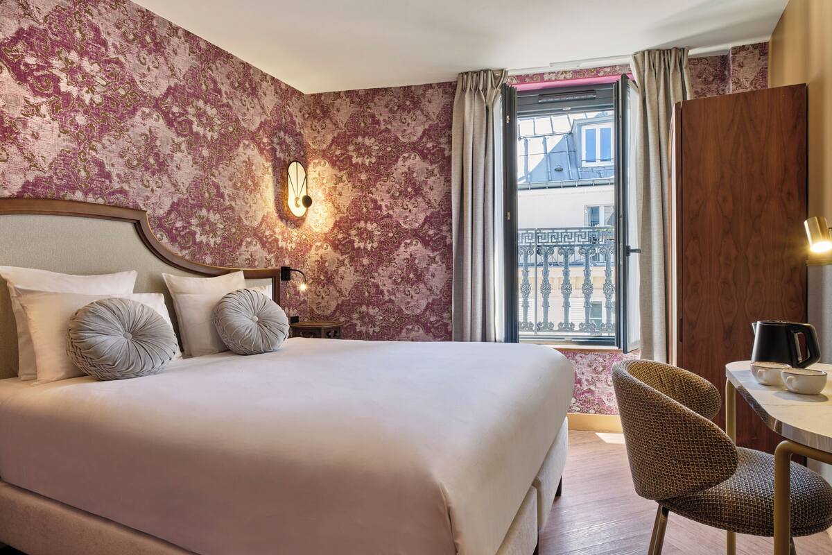 Habitación de hotel ordenada con una cama grande, una mesita con un hervidor y una silla.