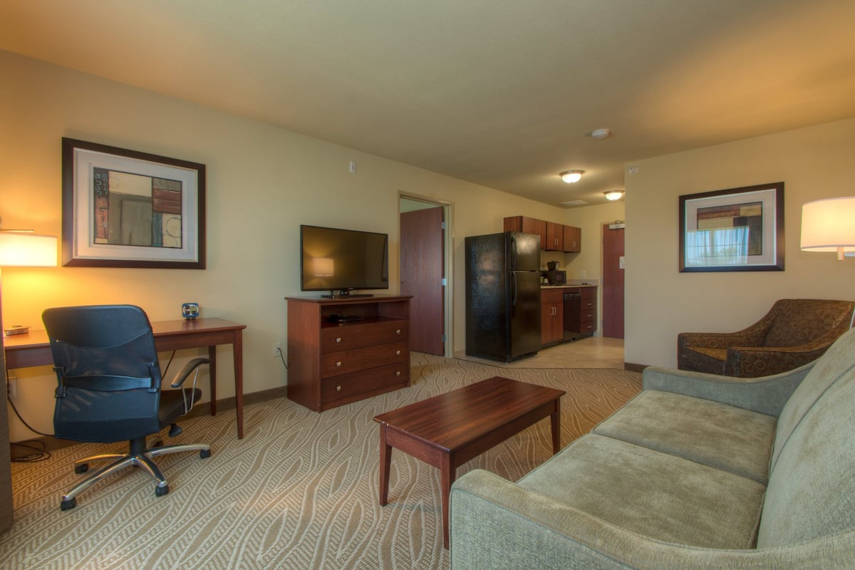 A hotel room with a sofa, a desk with a computer, a television, a refrigerator, and a kitchen area.