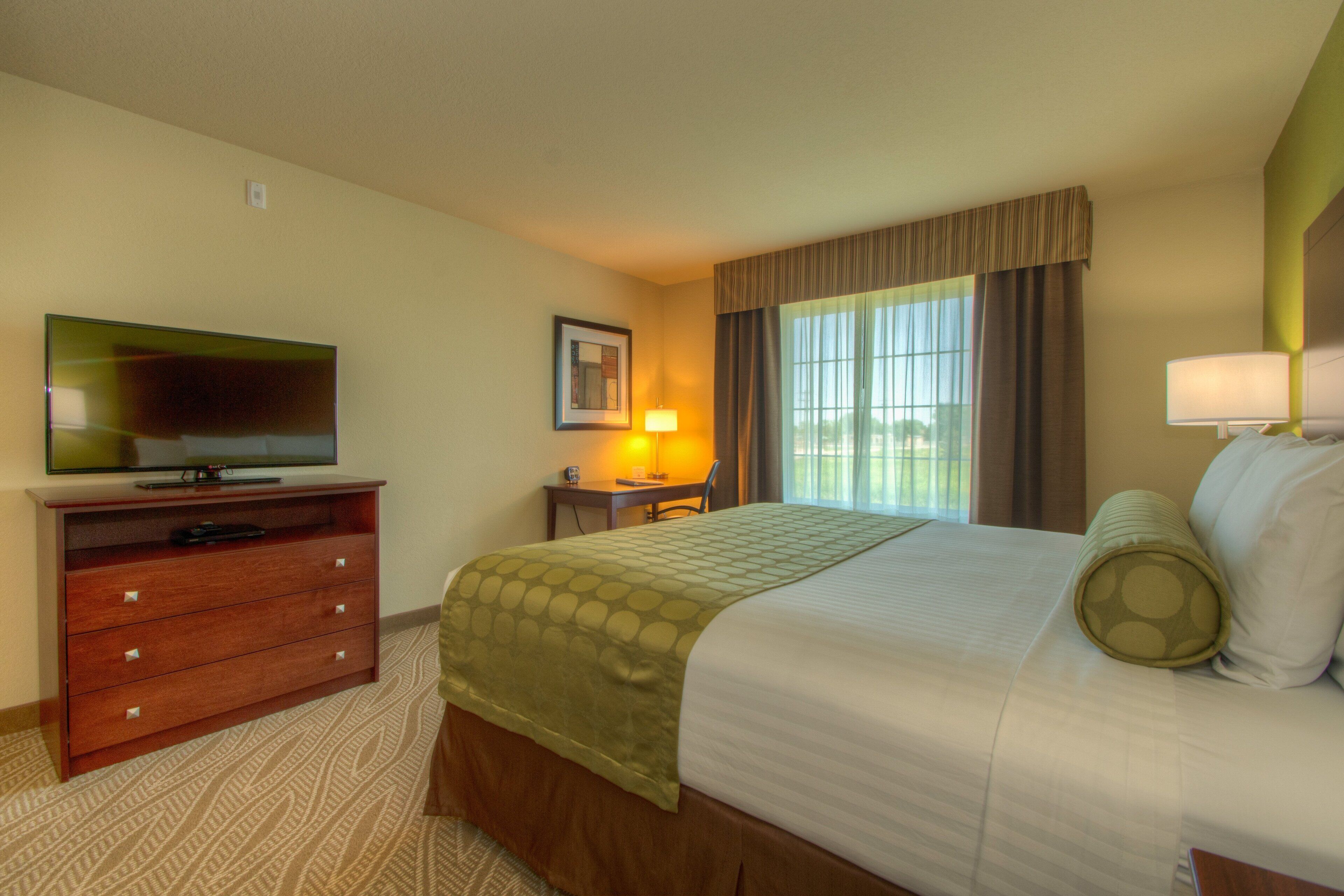 A hotel room with a bed, a television on a wooden dresser, a desk with a lamp, and a window with curtains.