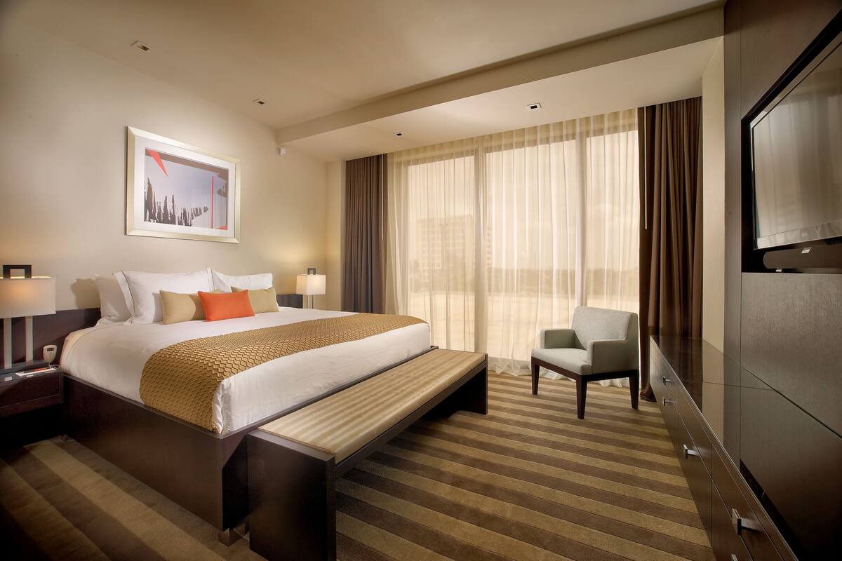 A hotel room with a large bed, a bench, a chair, a TV, and a framed picture on the wall.