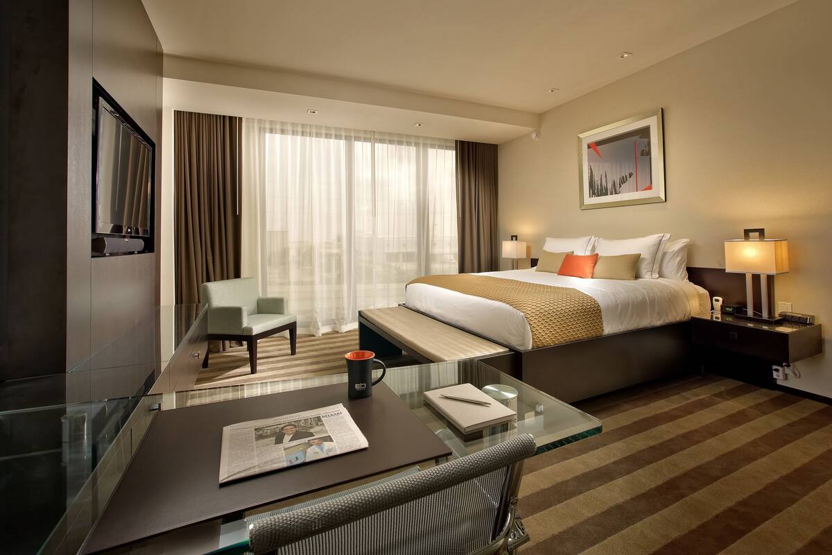 A hotel room with a bed, bedside tables, a desk, a chair, a TV, and a large window with curtains.
