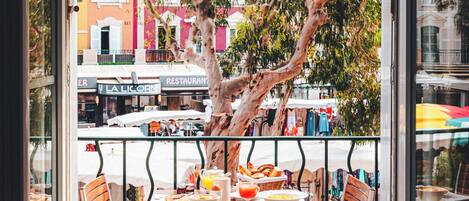 A balcony with a table set for breakfast, chairs, and a view of a street with buildings and trees.