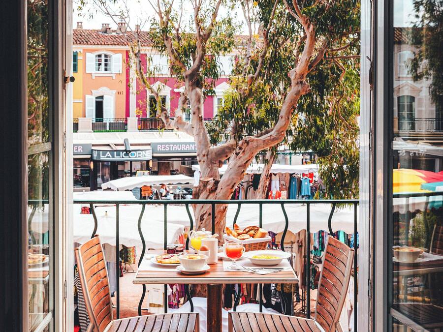 Een balkon met een ontbijttafel, stoelen en uitzicht op een straat met gebouwen en bomen.