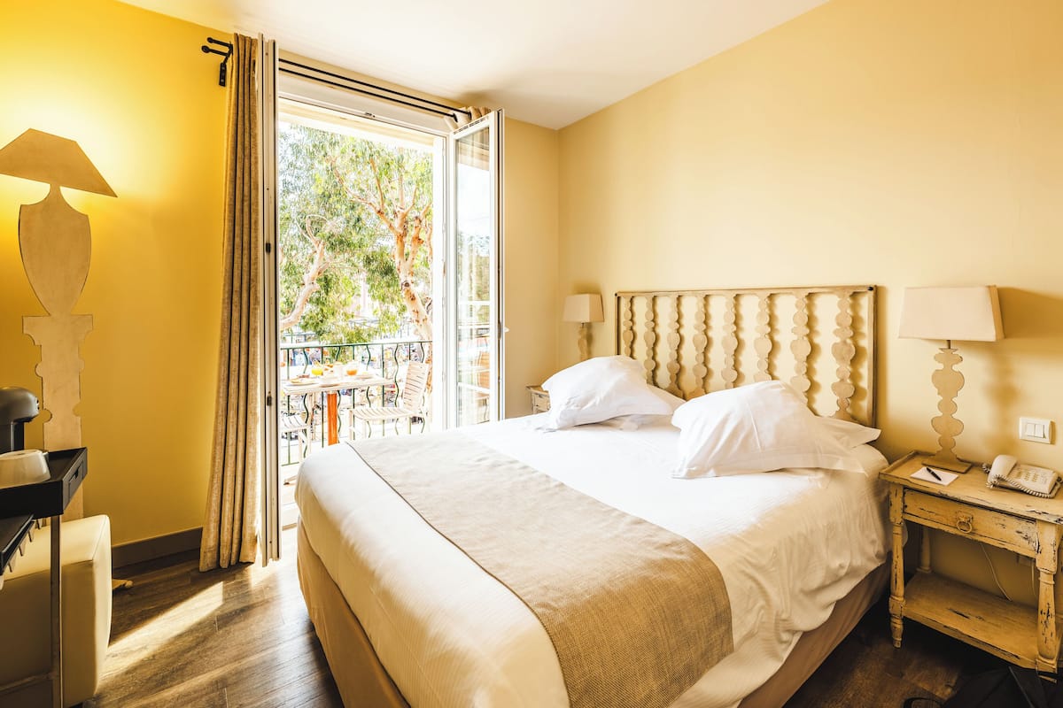 A hotel room with a large bed, a bedside table with a lamp, a nightstand with a phone, and a view of the outdoors through an open window.