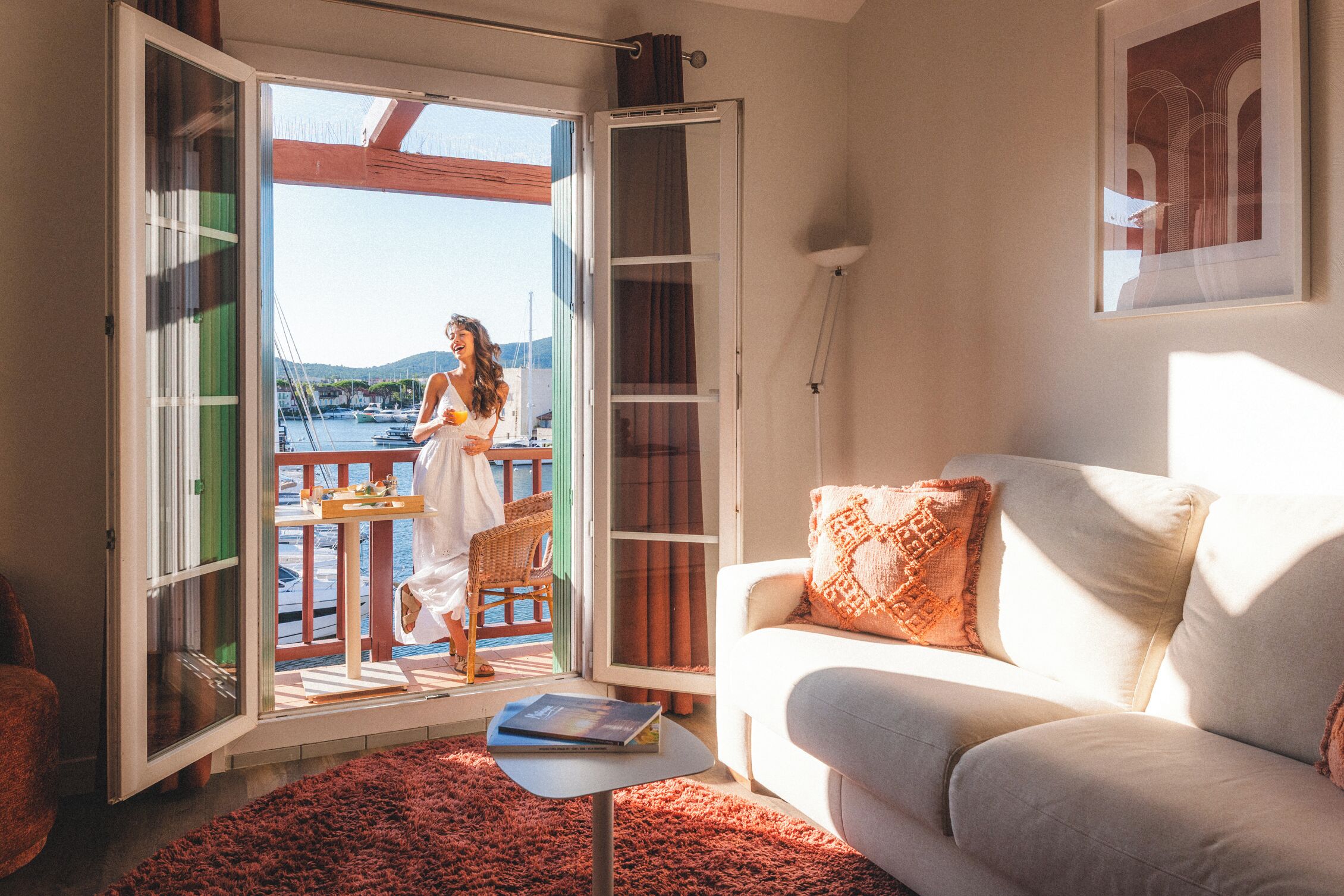 Una stanza con un divano bianco, un tappeto rosso e un balcone con vista su barche e colline.