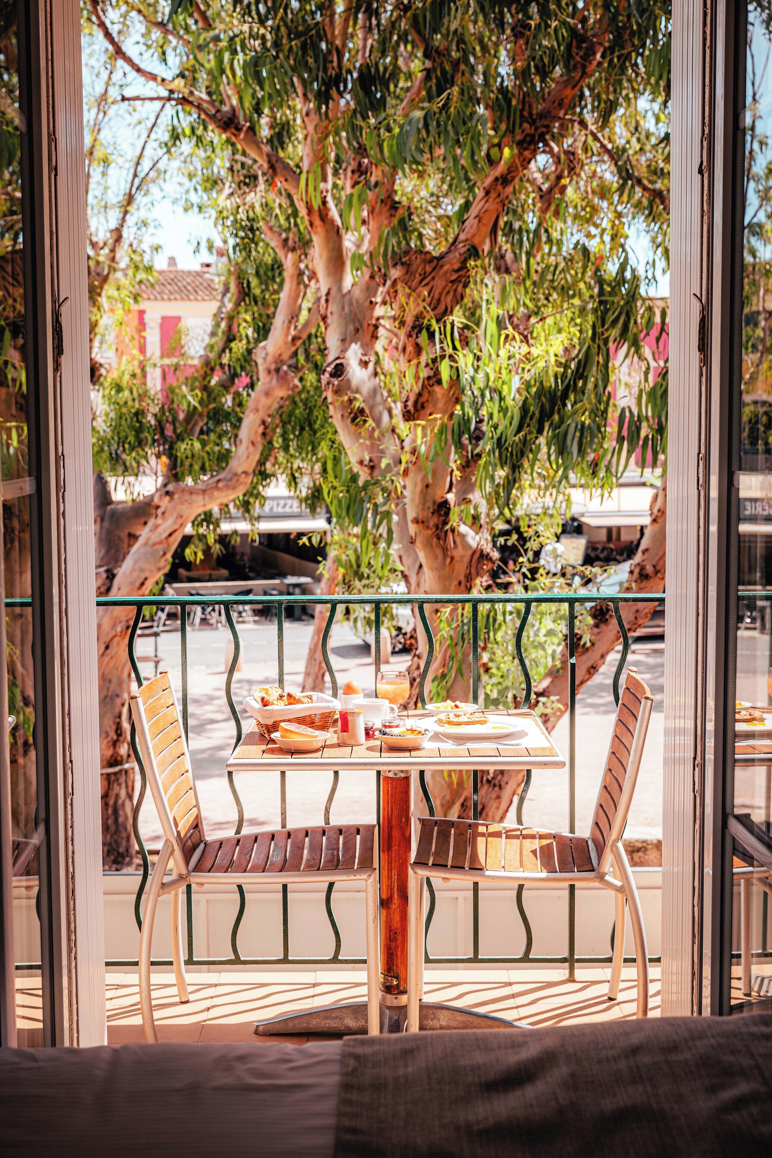 Un balcón con mesa y sillas, con vista a una calle con coches aparcados y un árbol.