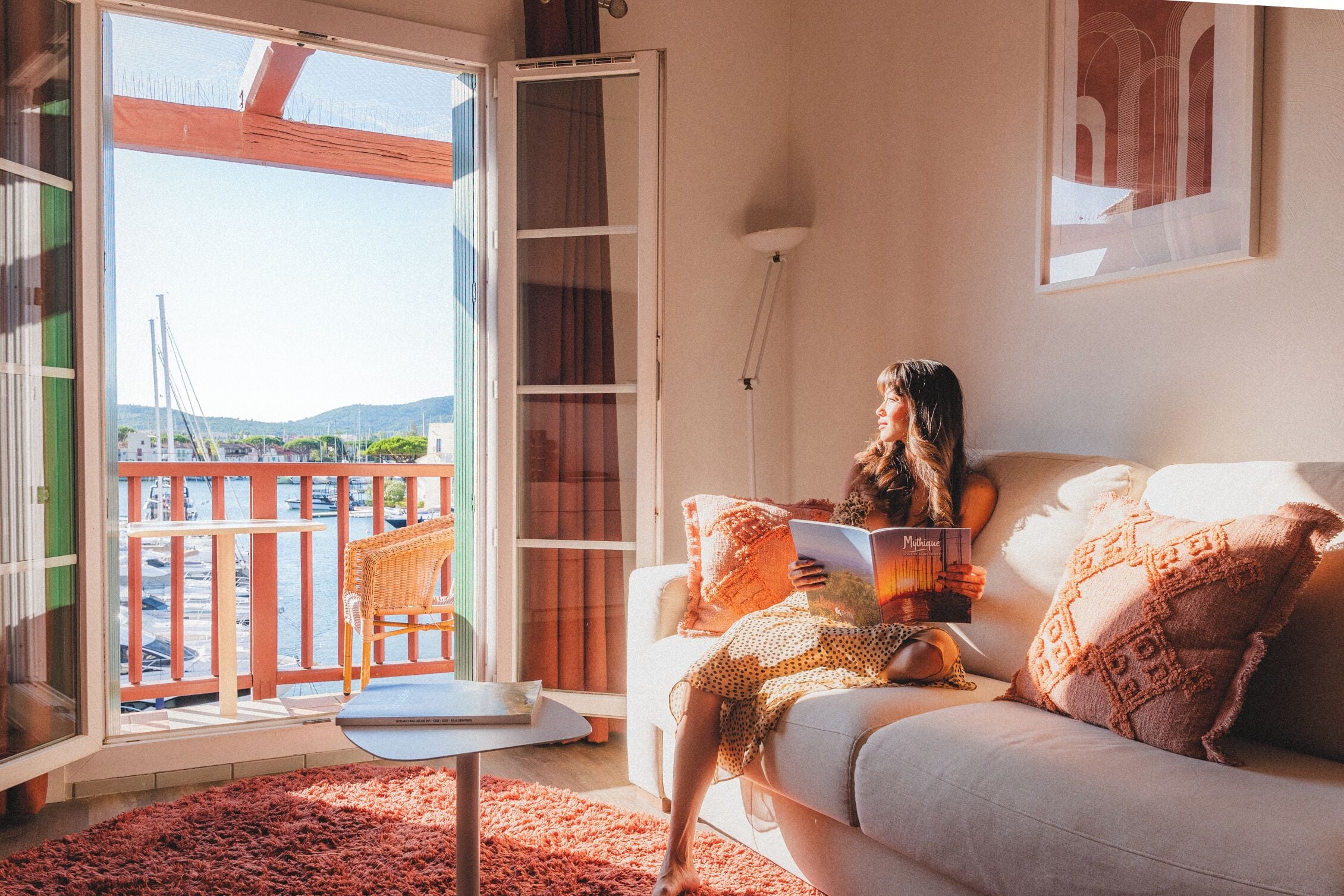 Uma sala de estar acolhedora com sofá, poltrona de leitura e vista para uma varanda com guarda-corpo e mesa.