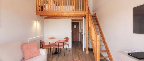 Un espacio tipo loft con una escalera de madera, un sofá blanco, una mesa de comedor con sillas y una pequeña kitchenette.