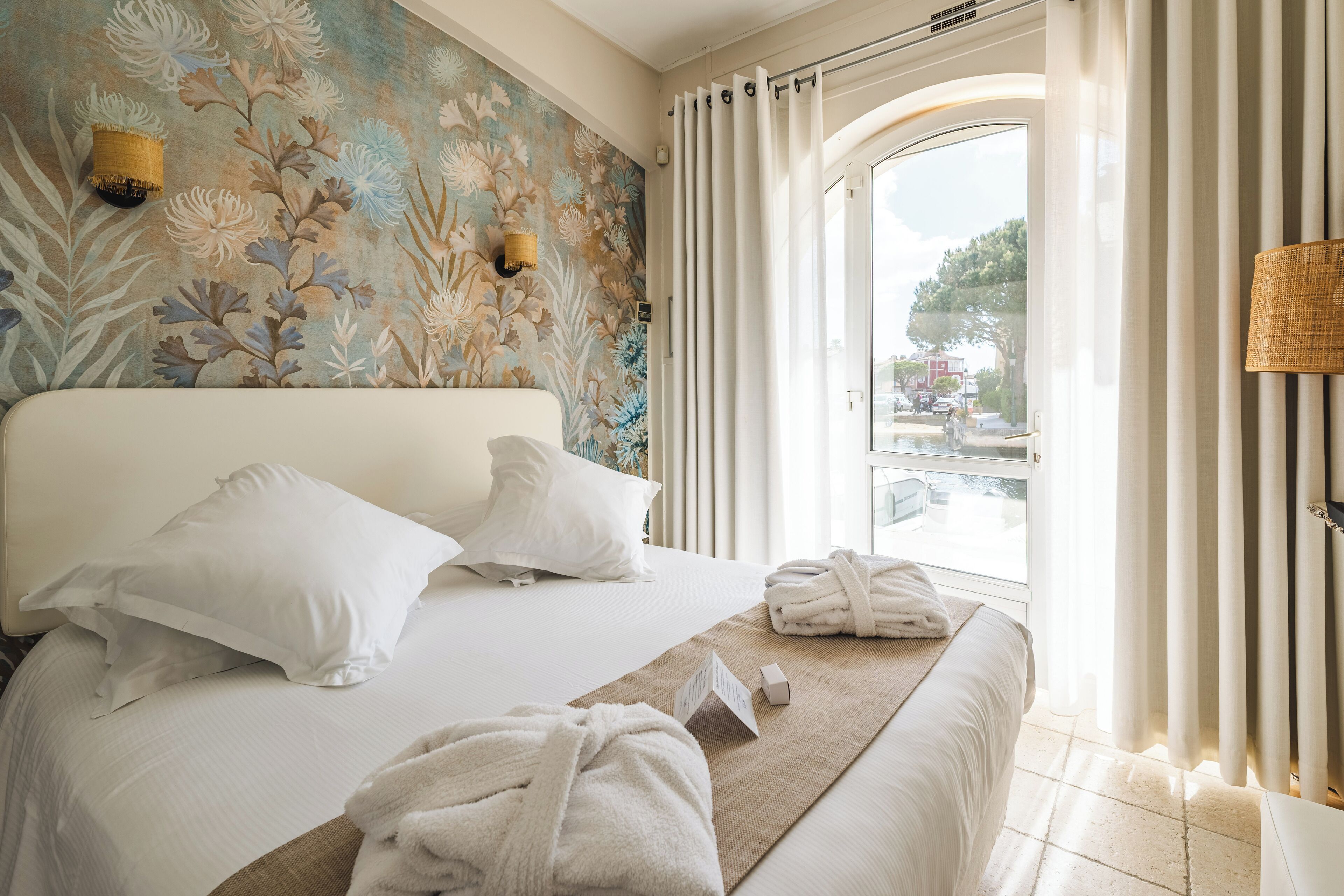 Habitación de hotel con una cama grande, dos almohadas, una mesita de noche con lámpara, una ventana con cortinas y un mural floral en la pared.