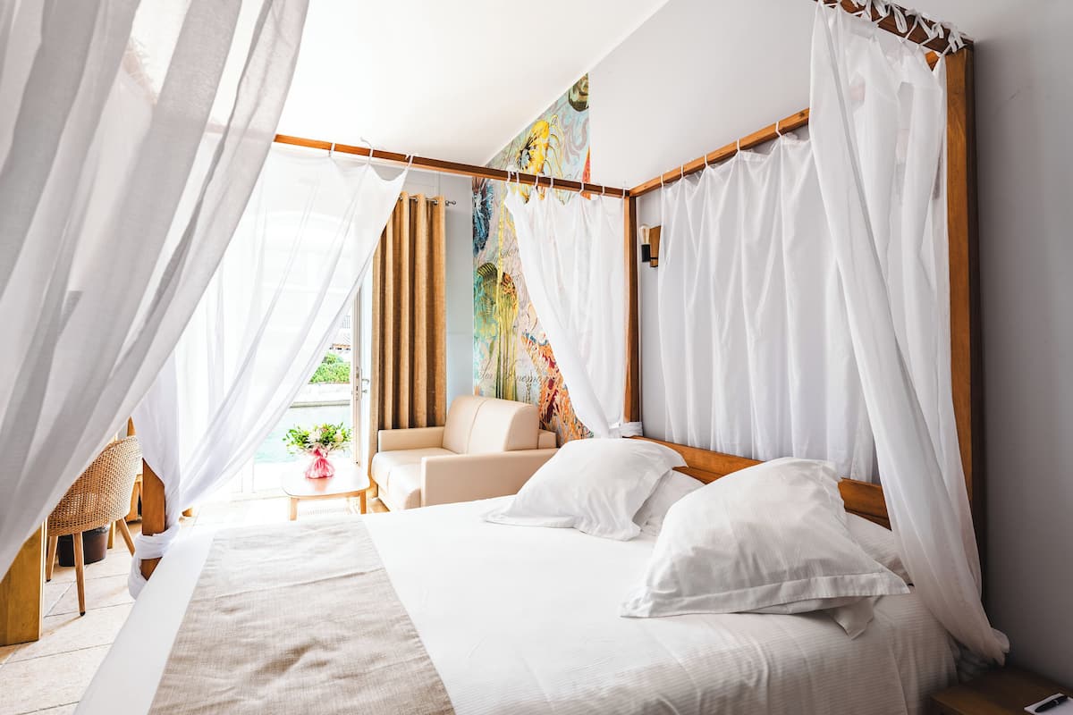 A bedroom with a bed, white curtains, a wooden headboard, a small table with a plant, and a colorful mural on the wall.