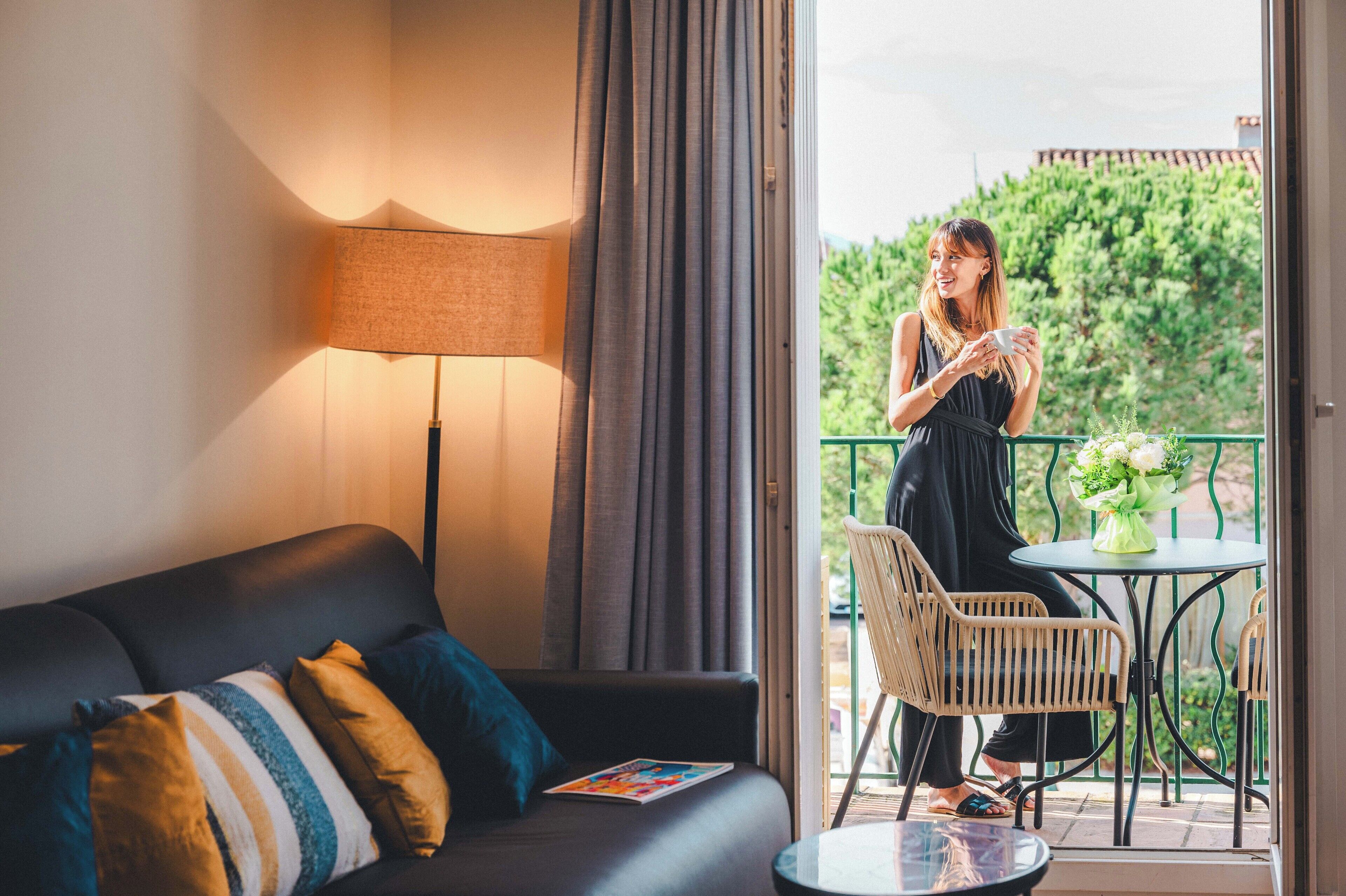 A room with a sofa, a table, a lamp, and a balcony with a view of greenery.