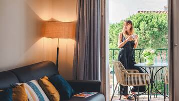A room with a sofa, a table, a lamp, and a balcony with a view of greenery.