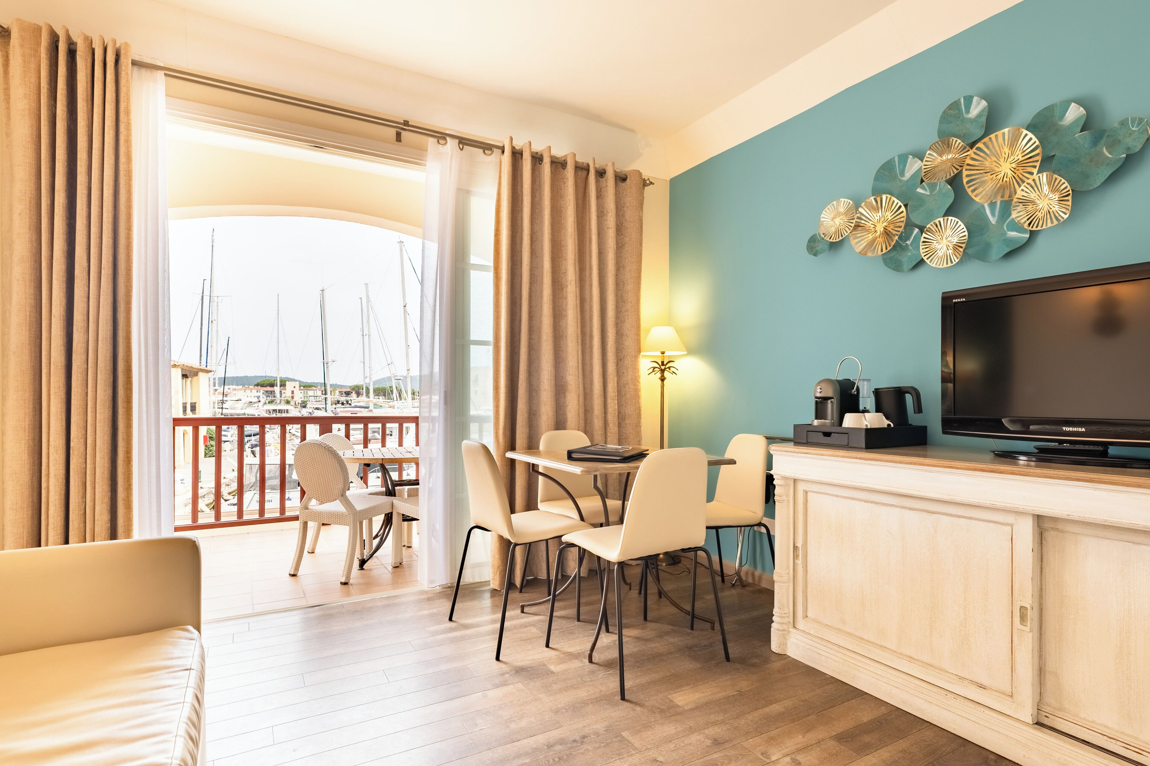 A hotel room with a balcony, a dining area with chairs and a table, a TV, and a wall with decorative leaves.