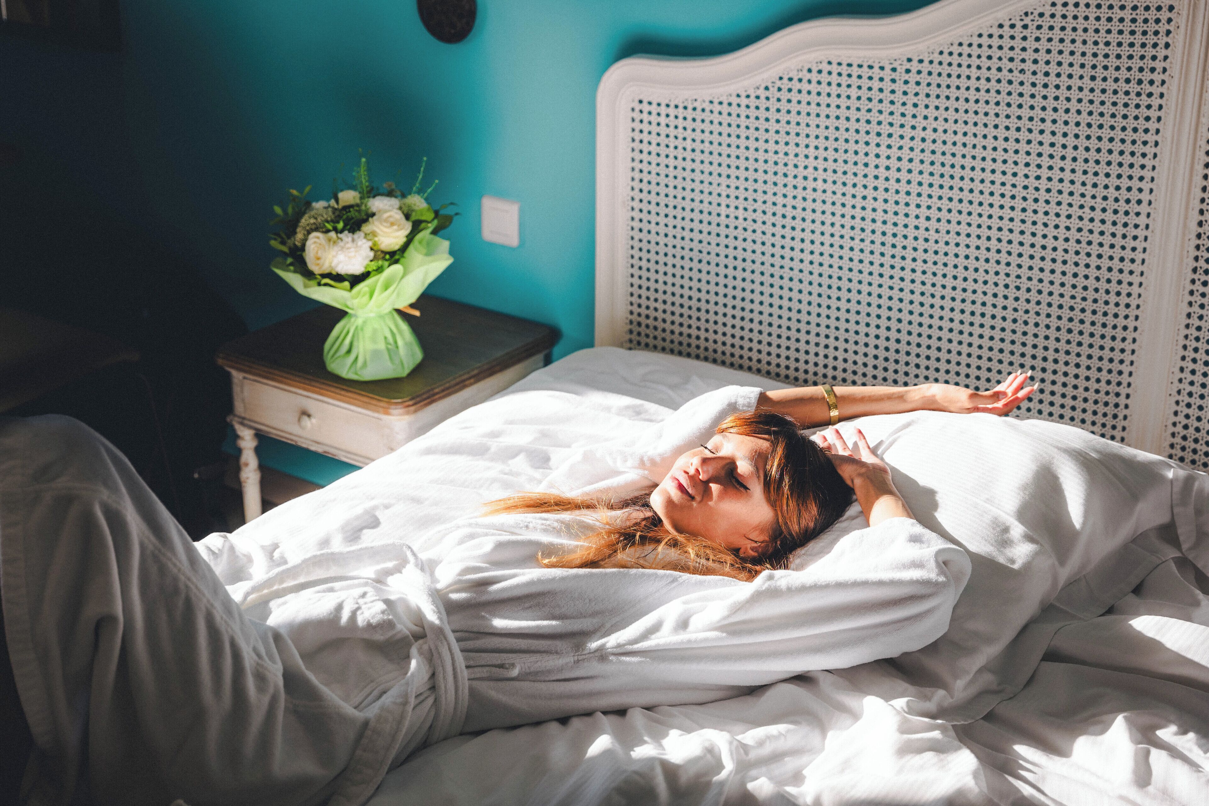 Una cama con ropa de cama blanca, una mesita de noche con un florero y una pared azul.