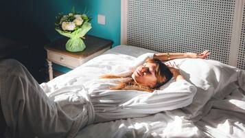 Una cama con ropa de cama blanca, una mesita de noche con un florero y una pared azul.
