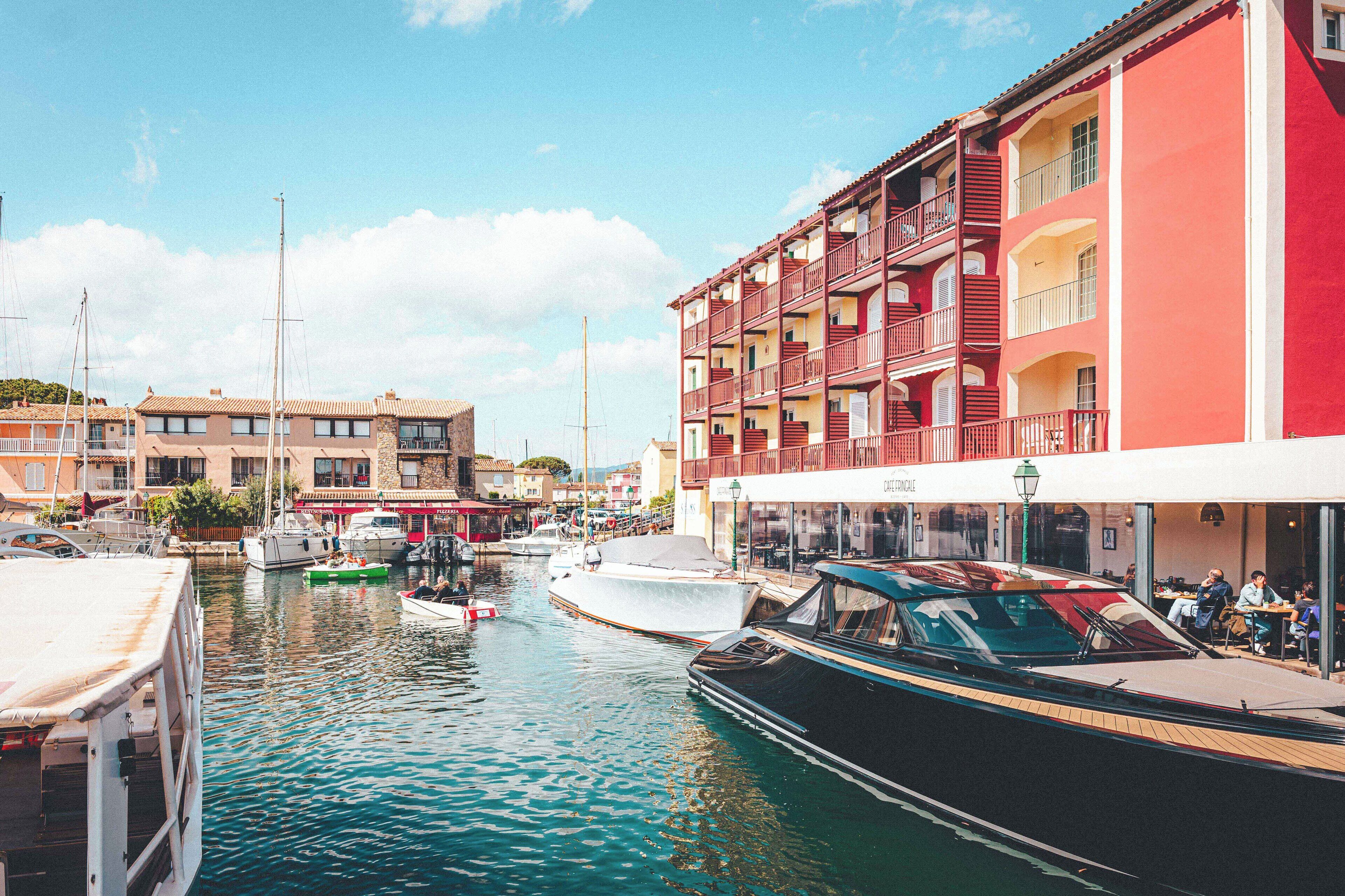 Un port de plaisance avec des bateaux amarrés, un immeuble doté de balcons et un café où des gens sont assis à l’extérieur.