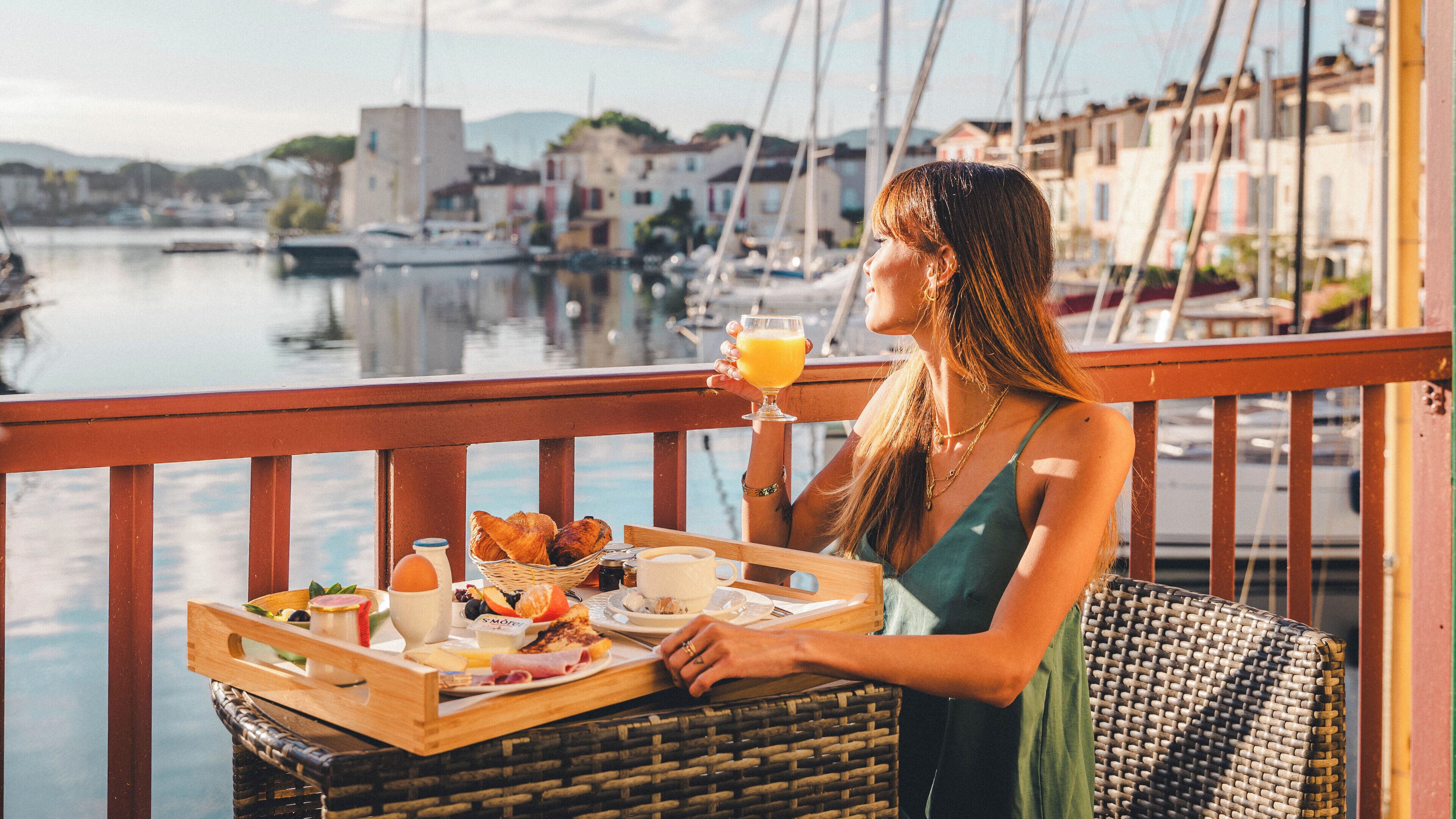 Una mujer disfrutando del desayuno en un balcón con vista a un puerto deportivo y edificios.