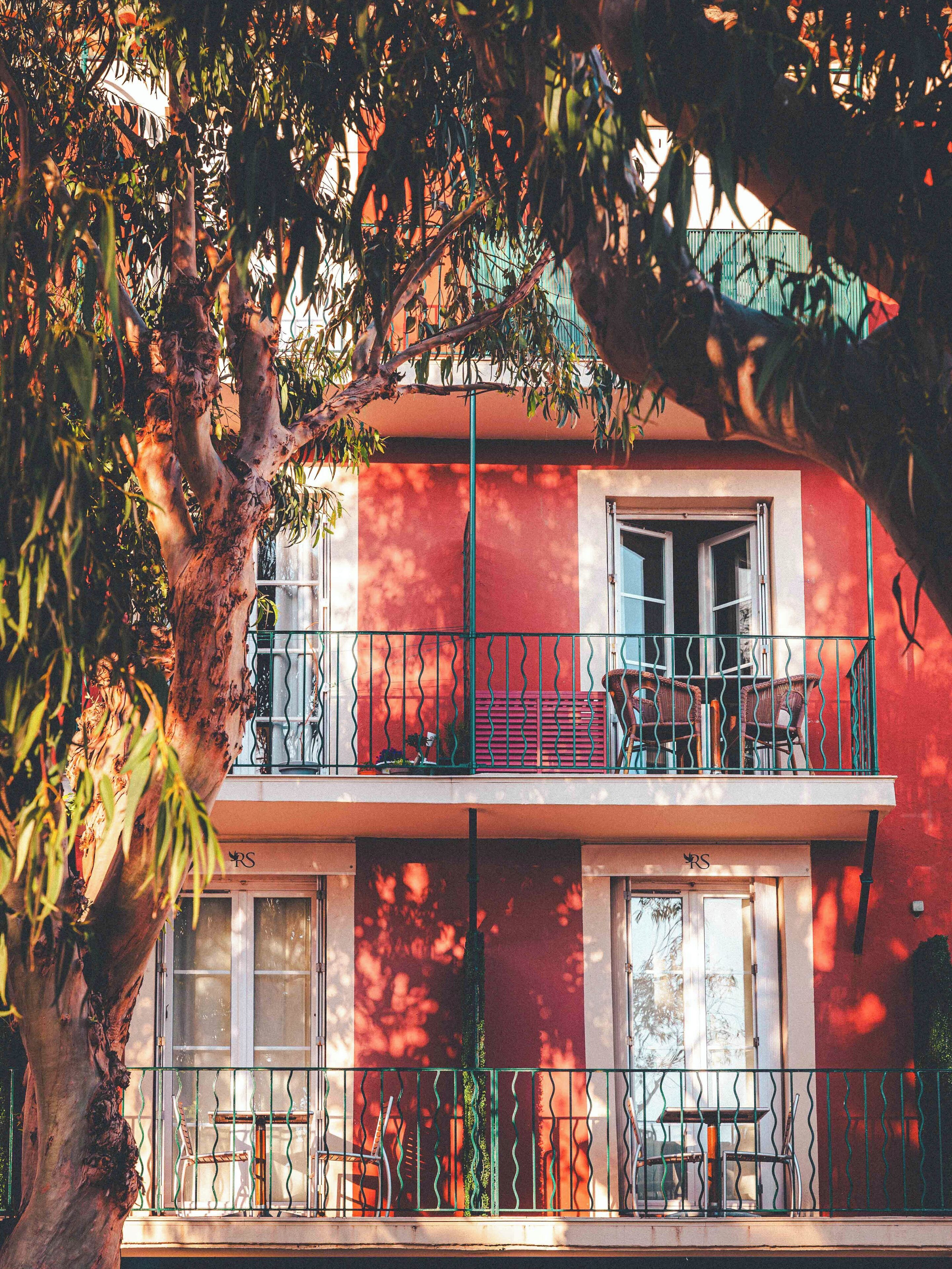 A two-story building with a balcony, a tree in the foreground, and a red wall.