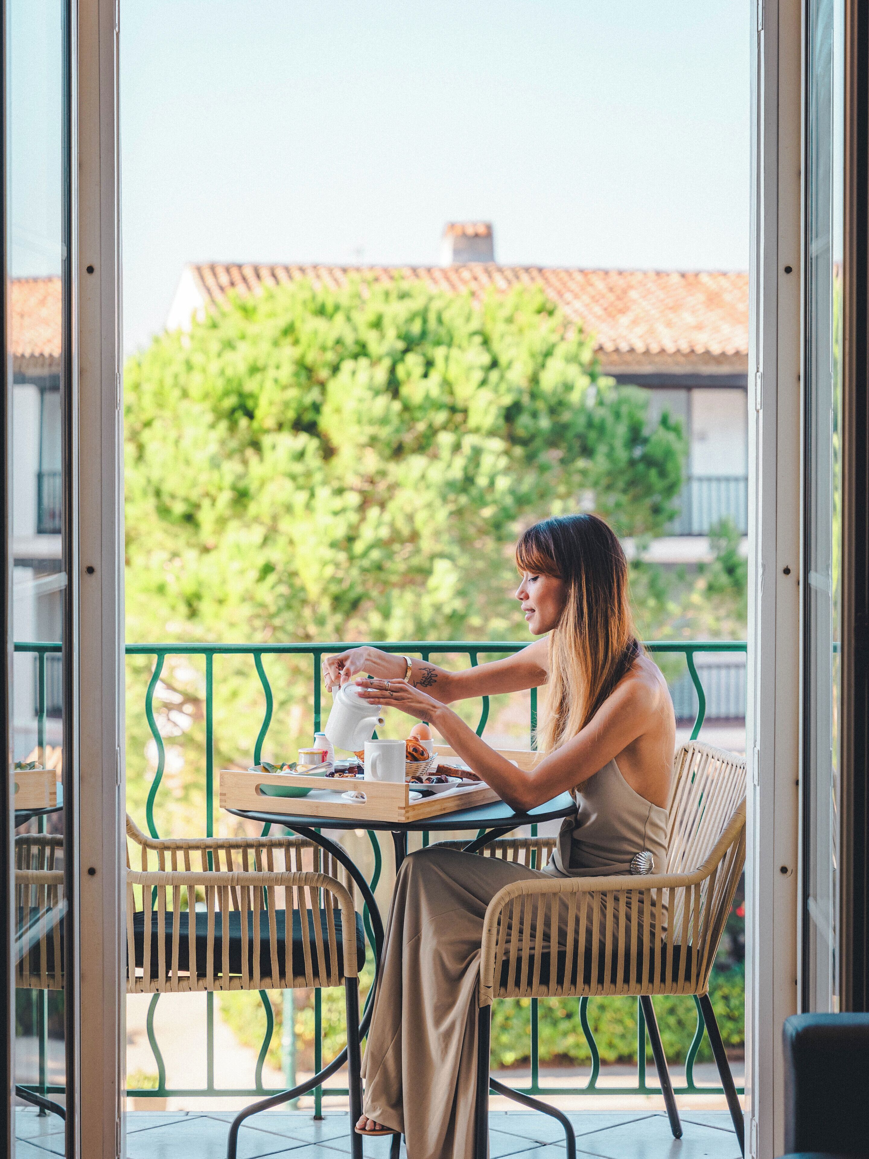 Iemand zit aan een tafel met een dienblad eten en een theepot, op een balkon met een groene reling en uitzicht op bomen en gebouwen.