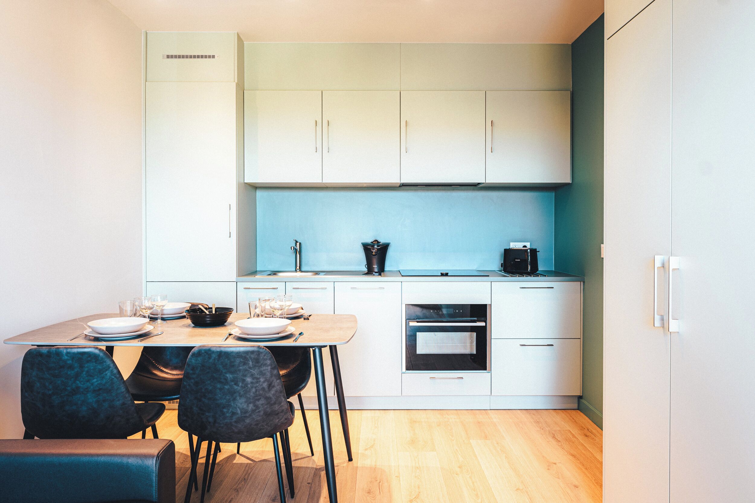 Una cocina moderna con armarios blancos, un salpicadero azul y una zona de comedor con mesa de madera y sillas oscuras.