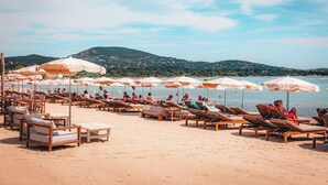 A beach with rows of sun loungers and umbrellas, a sandy beach, and a hilly landscape in the background. -  Le Suffren Hotel, Golfe de Saint-Tropez (Grimaud)