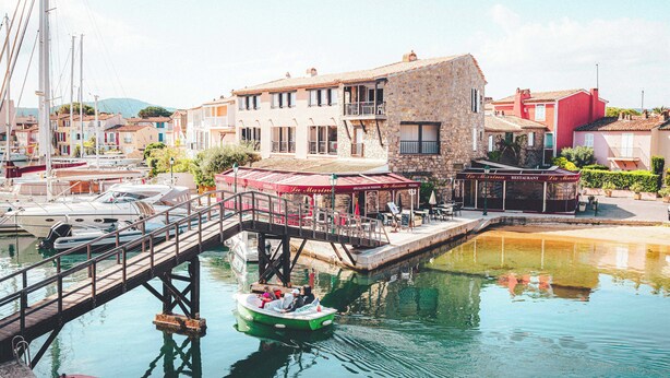 A marina with boats, a wooden bridge, and buildings with outdoor seating. -  Le Suffren Hotel, Golfe de Saint-Tropez (Grimaud)