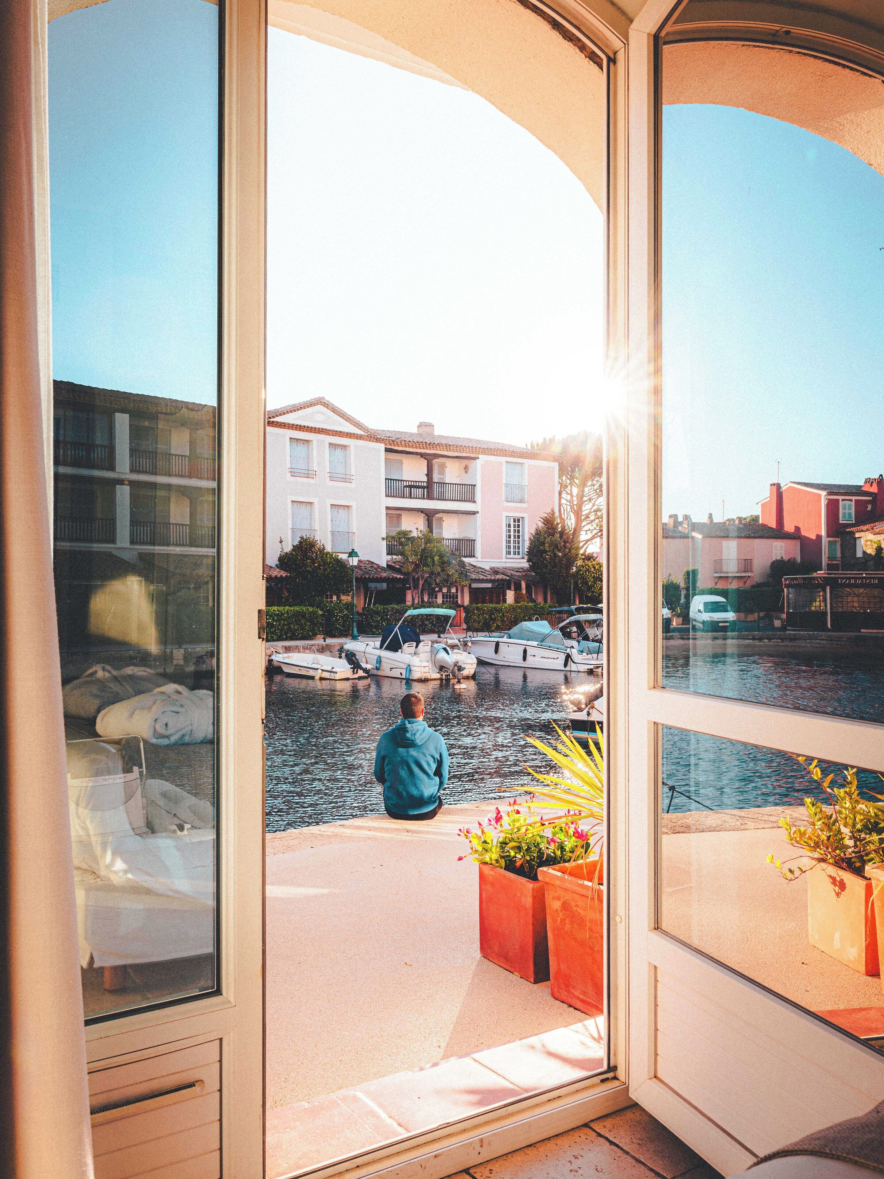A person sitting by a window overlooking a marina with boats and buildings in the background.