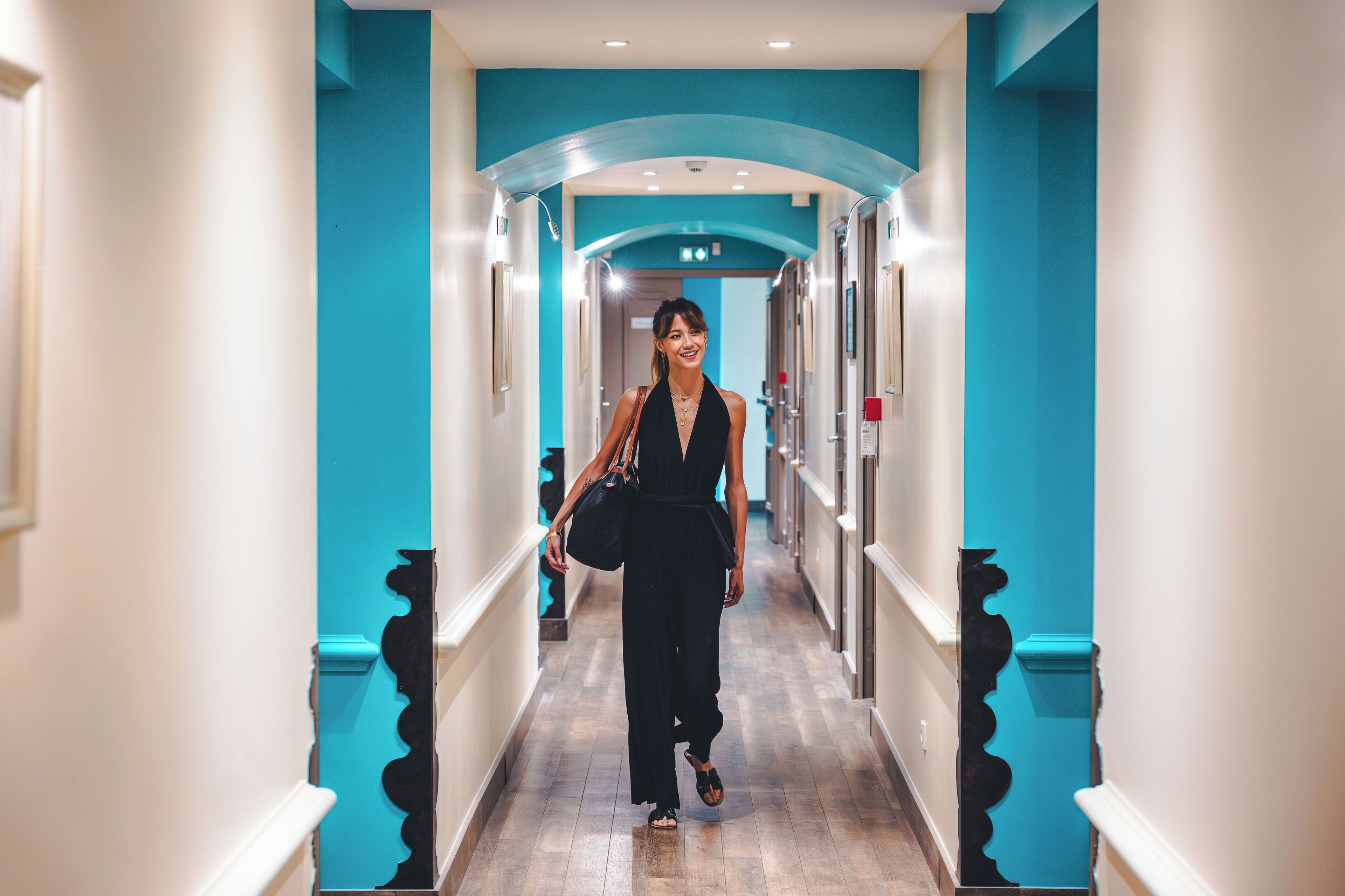 Un pasillo de hotel con decoración en tonos aguamarina, una mujer caminando y un diseño moderno.