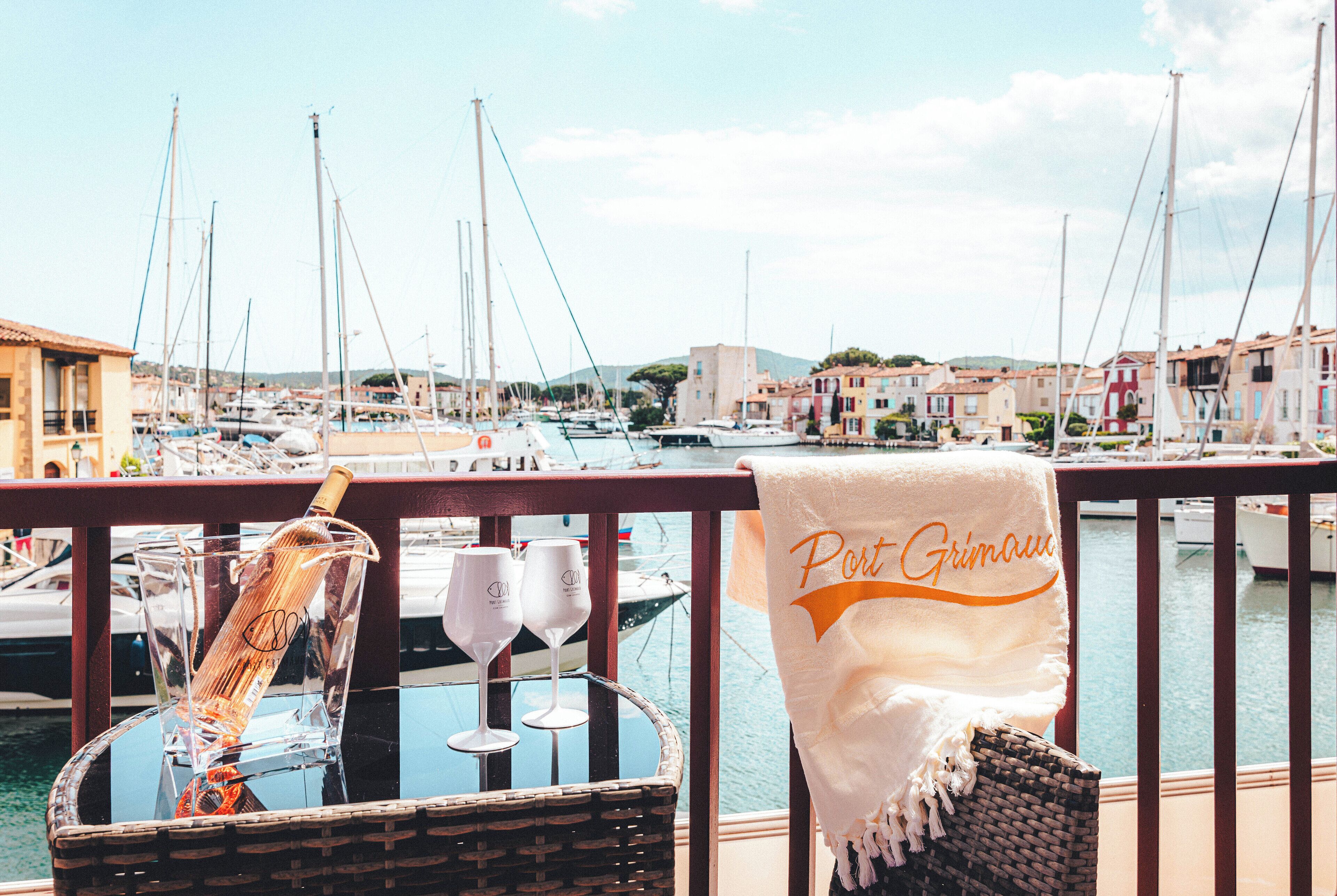 A balcony with a wicker chair, a bottle, and two wine glasses. A towel with "Port Grimaldi" is draped over the railing.