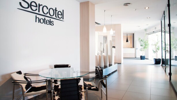 A modern hotel lobby with a glass table and chairs, a visible reception area, and a sign for 'Sercotel hotels'.