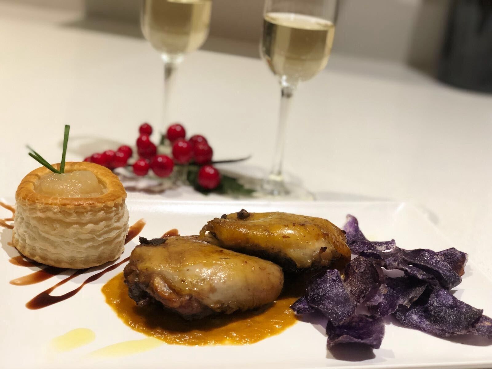 A dessert with a pastry on a plate, accompanied by two pieces of meat and purple vegetables, with two glasses of a clear liquid in the background.