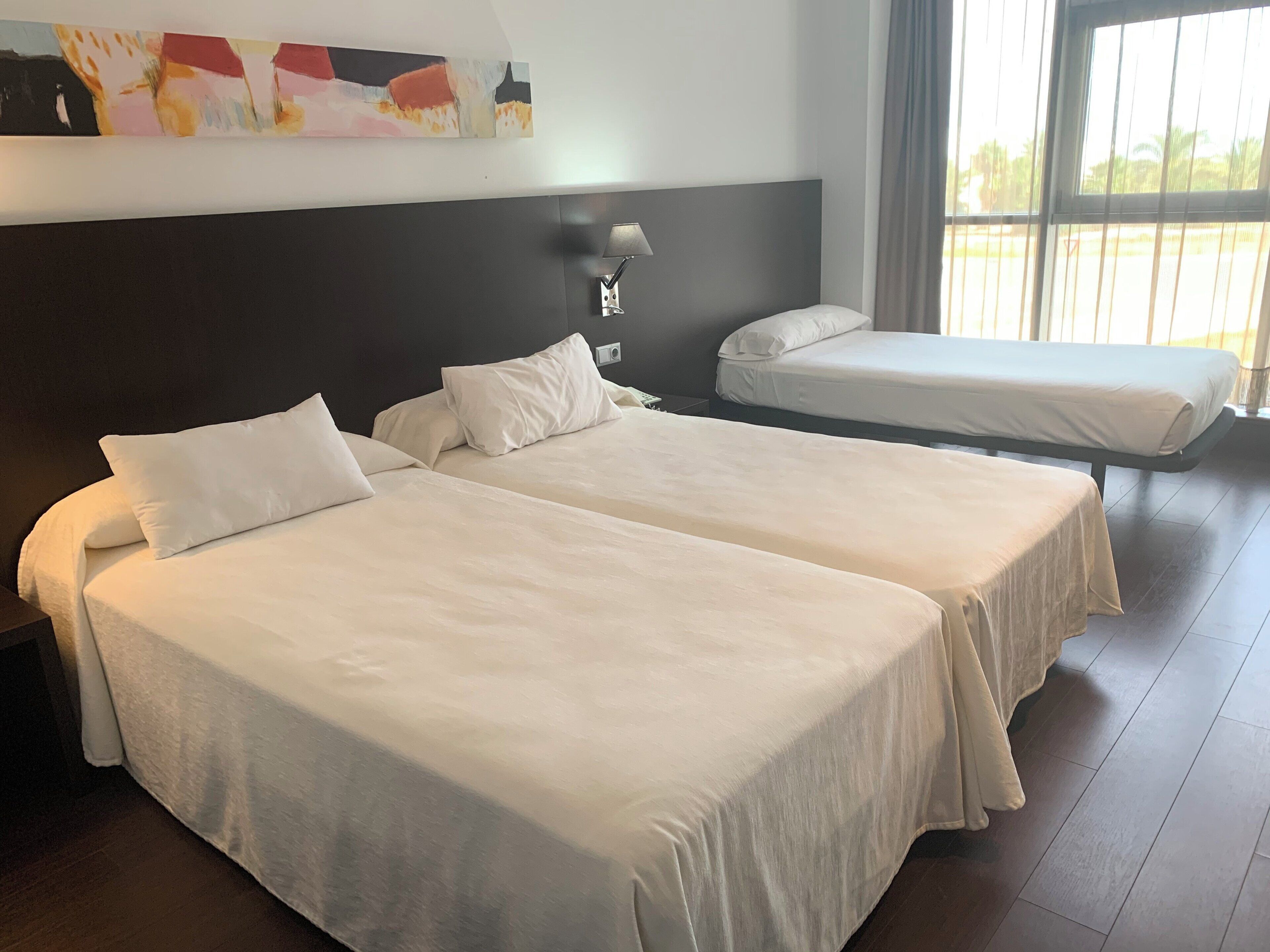 A hotel room with two beds, a nightstand, a wall-mounted lamp, and a large window with curtains.