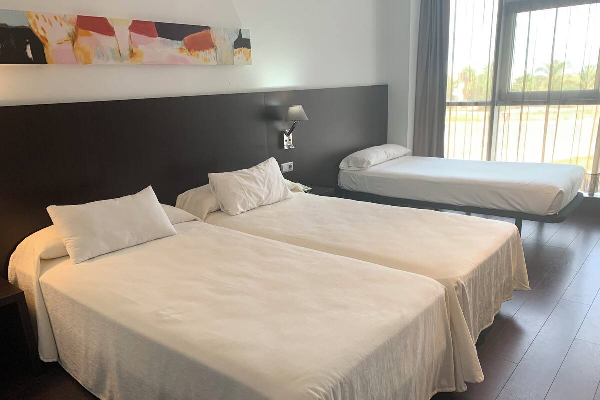 Een hotelkamer met twee bedden, een nachtkastje, een wandlamp en een groot raam met gordijnen.