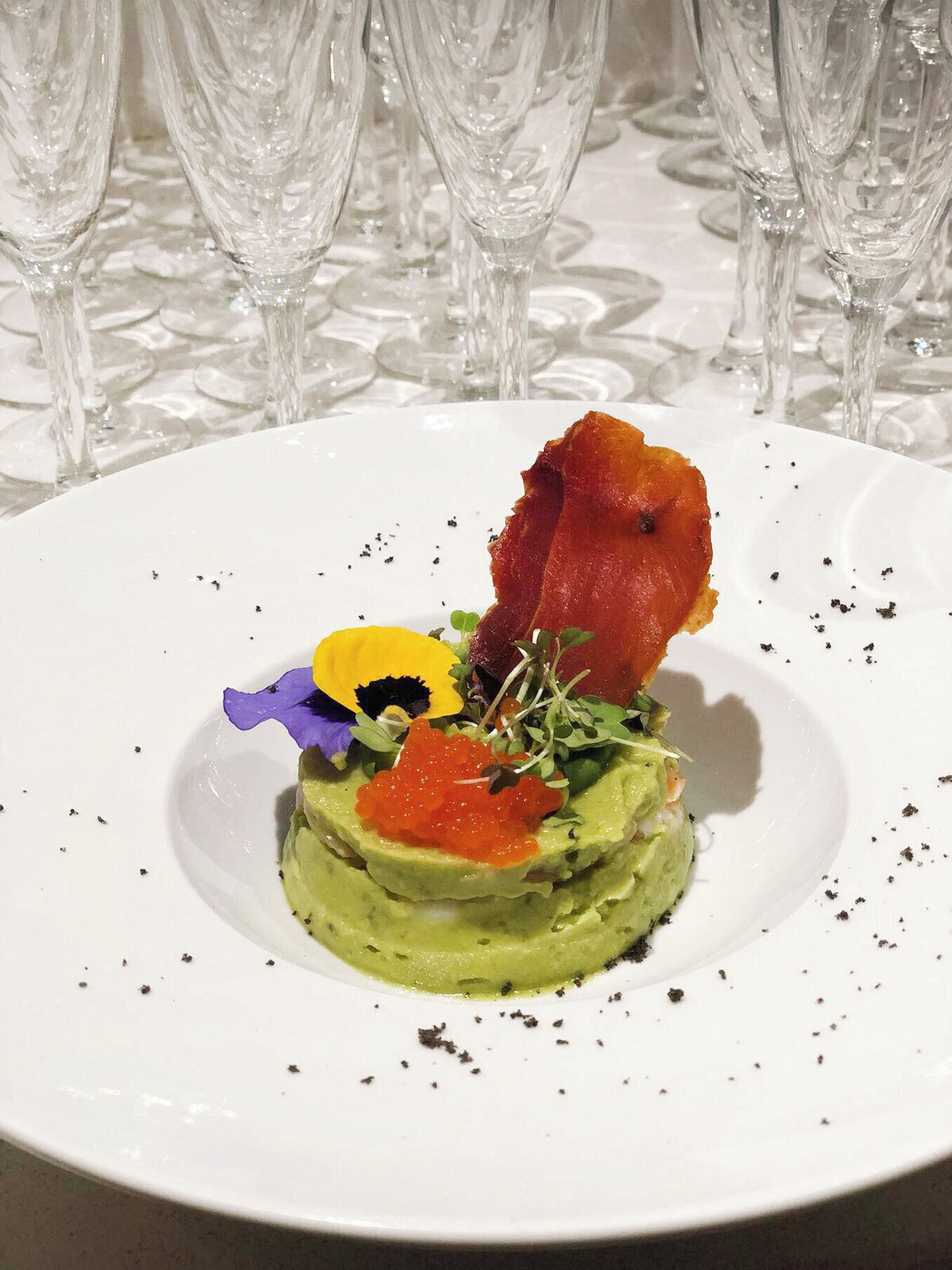 Un plato blanco con un plato a base de aguacate verde, adornado con caviar rojo, un trozo de tocino y flores comestibles.