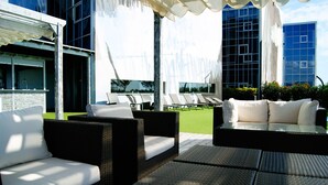 A rooftop terrace with wicker furniture, a pool, and modern buildings in the background.