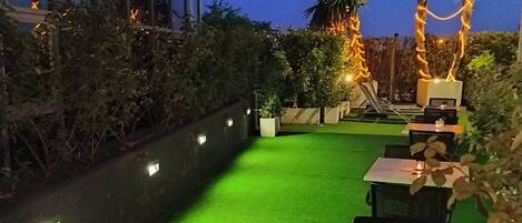 Un área de patio exterior con césped artificial verde, mesas y sillas.