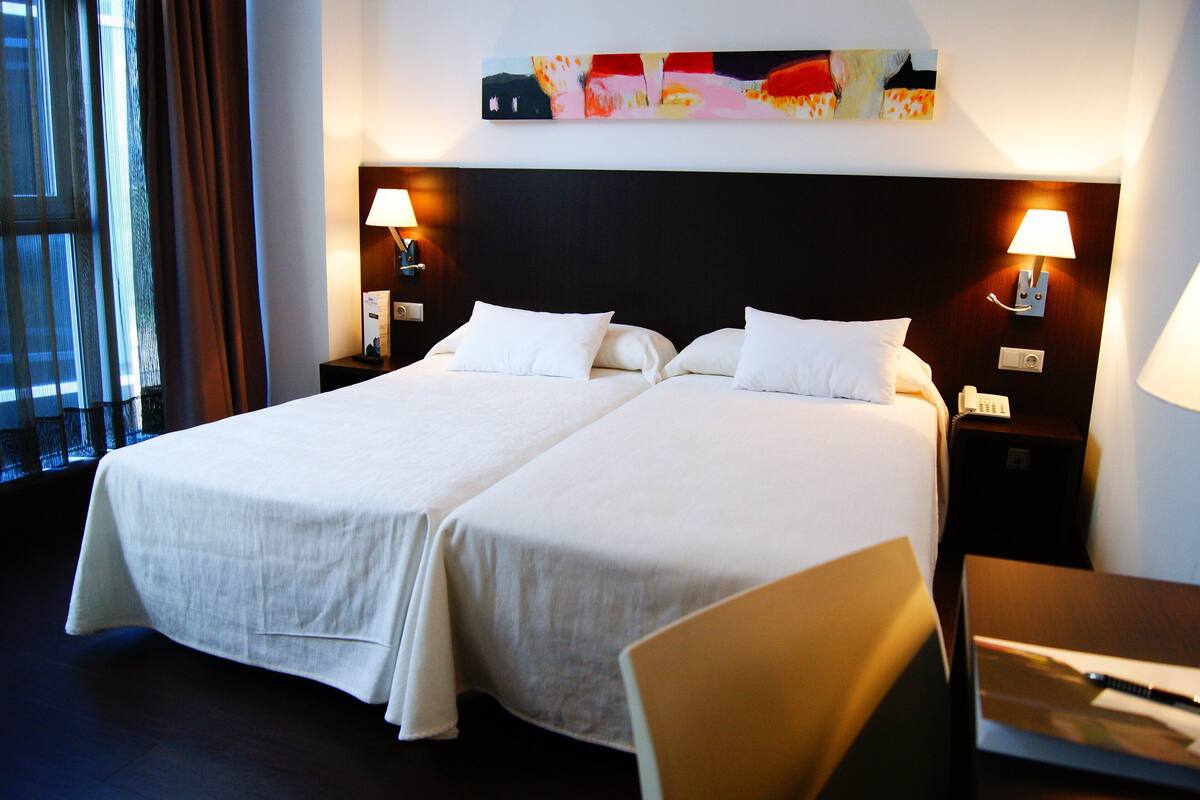 Une chambre d’hôtel avec deux lits, un bureau et une chaise. Il y a deux tables de chevet avec des lampes et un tableau au-dessus des lits.