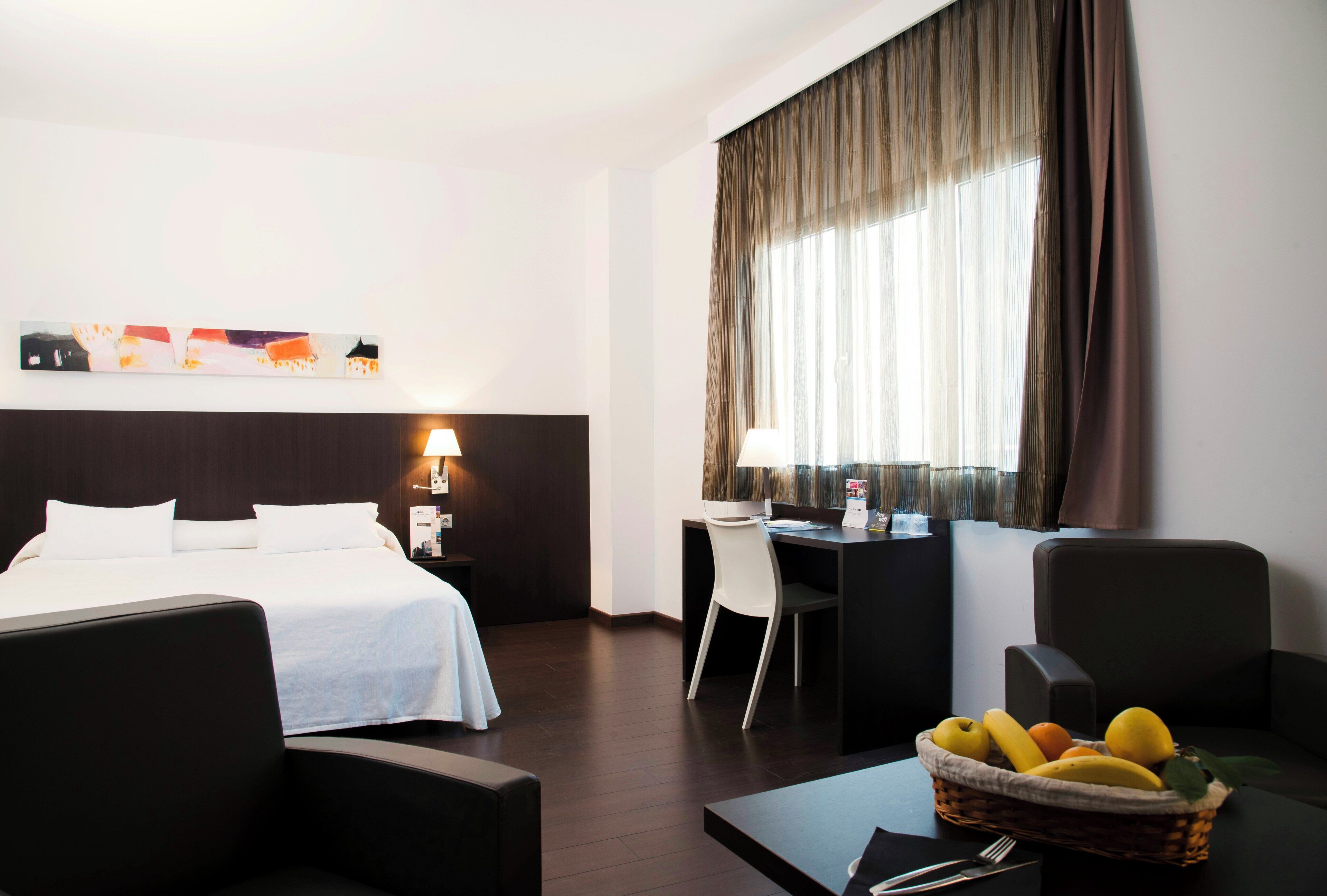 Una habitaci ón de hotel con una cama, escritorio, dos sillas, lámpara, cesta de frutas y un cuadro en la pared.