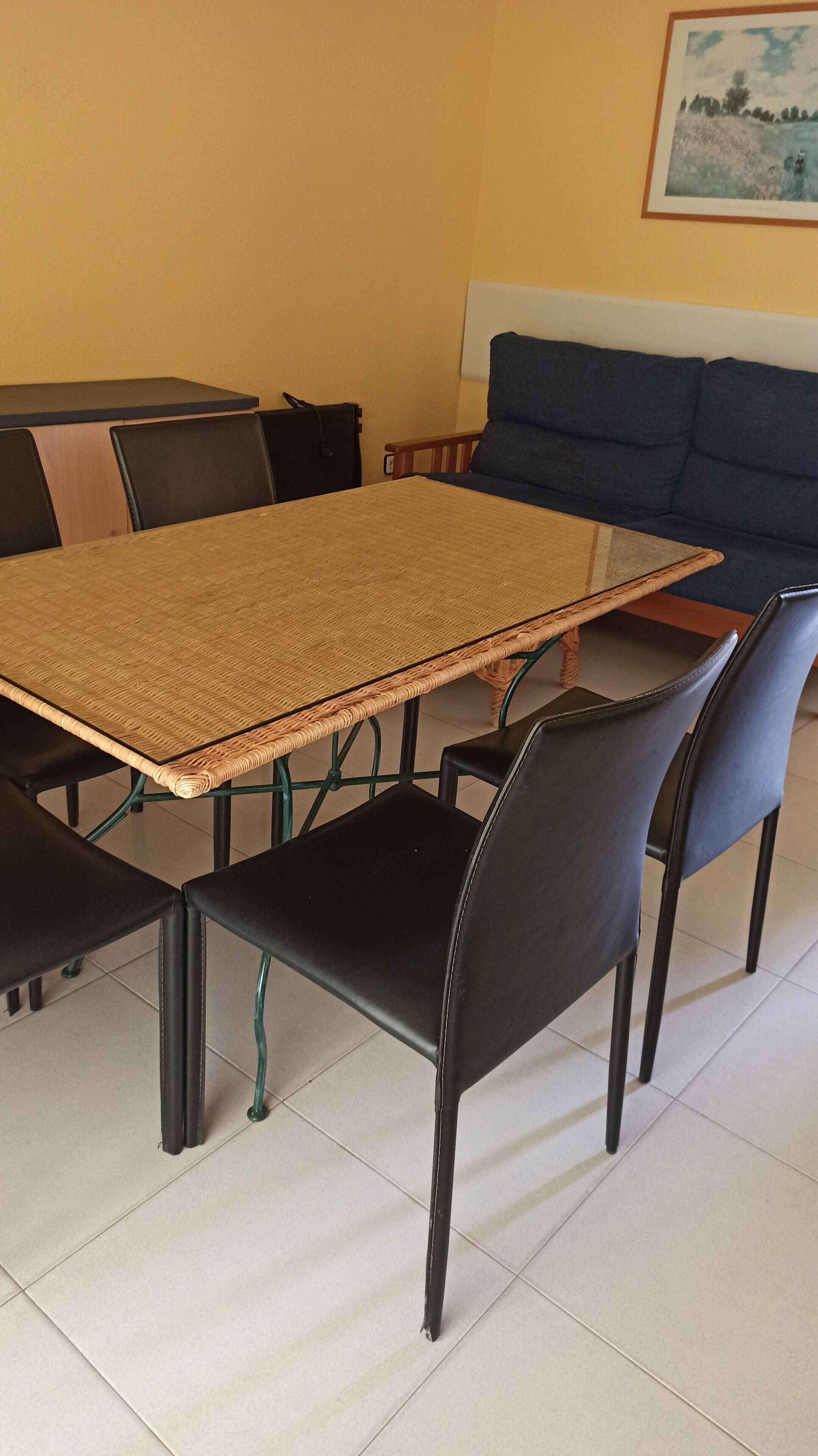 A dining area with a woven table and black chairs, a sofa, and a painting on the wall.