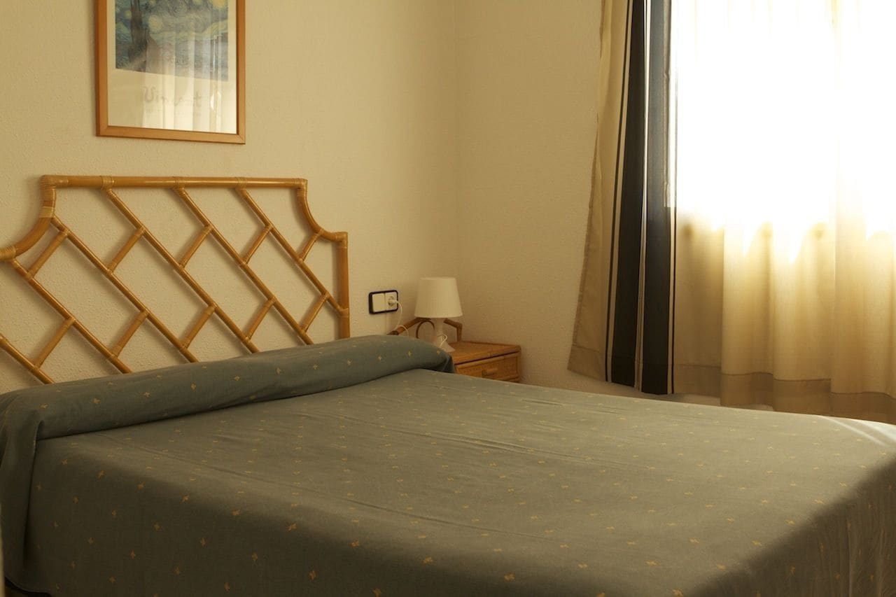 A neatly made bed with a patterned bedspread, a wooden headboard, a bedside table with a lamp, and a framed picture on the wall.