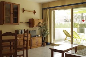Una sala de estar con un aparador de madera, televisor, planta y una puerta corrediza de vidrio que da a un balcón con sillas amarillas.