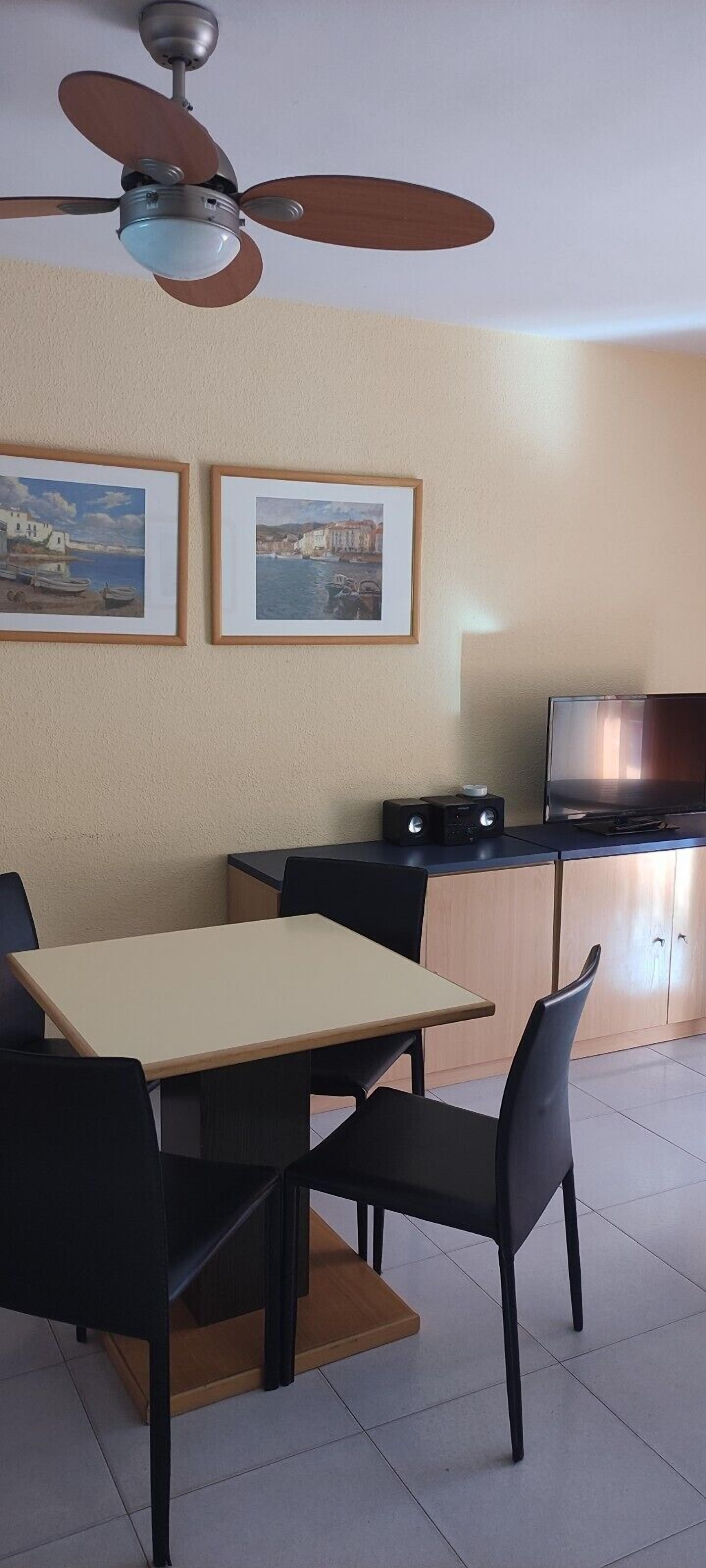 Un comedor con mesa y sillas, ventilador de techo, cuadros en la pared y un televisor sobre un mueble.