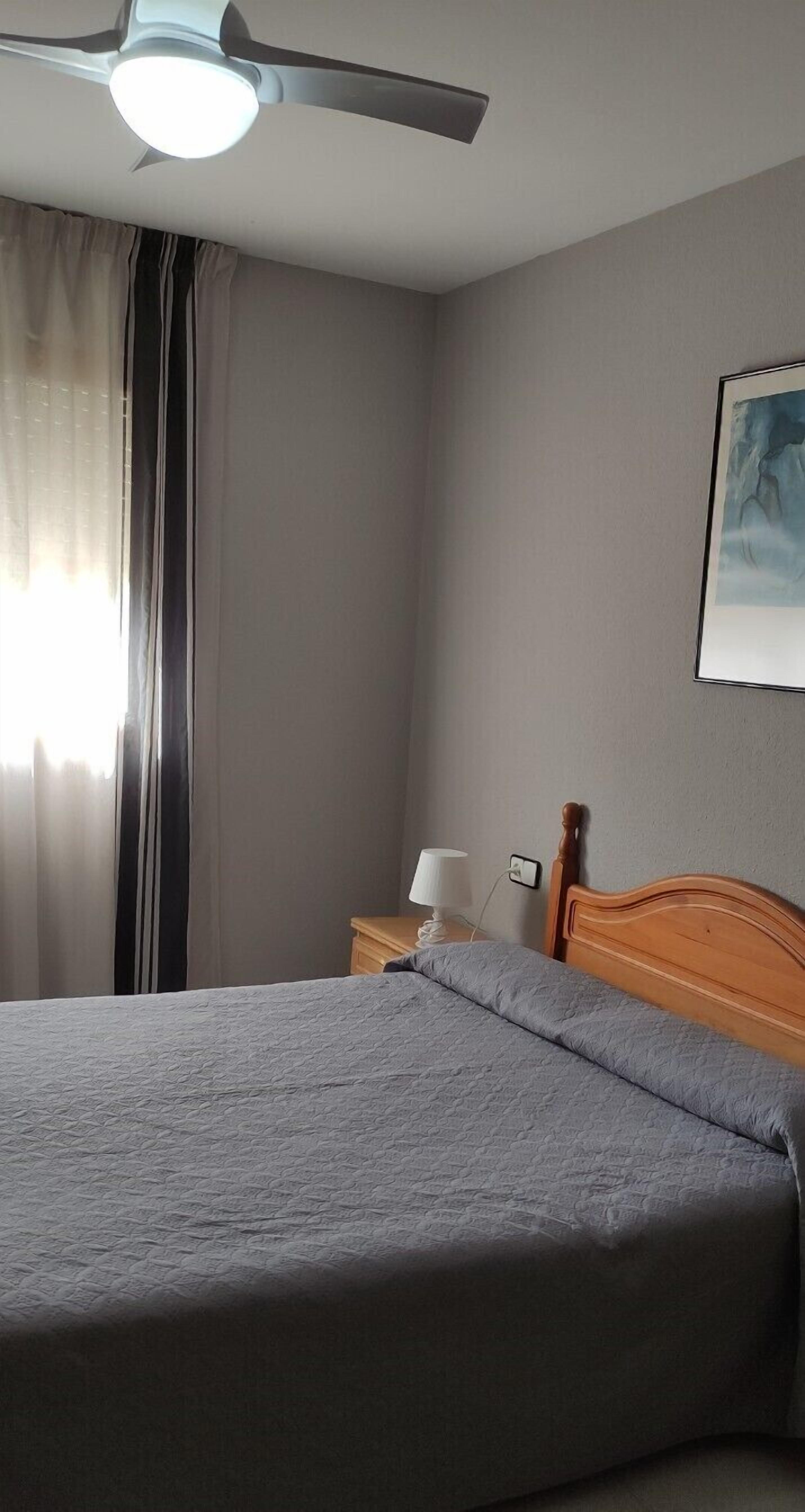 Un dormitorio con cama, mesita de noche, lámpara, ventilador de techo y un cuadro en la pared.