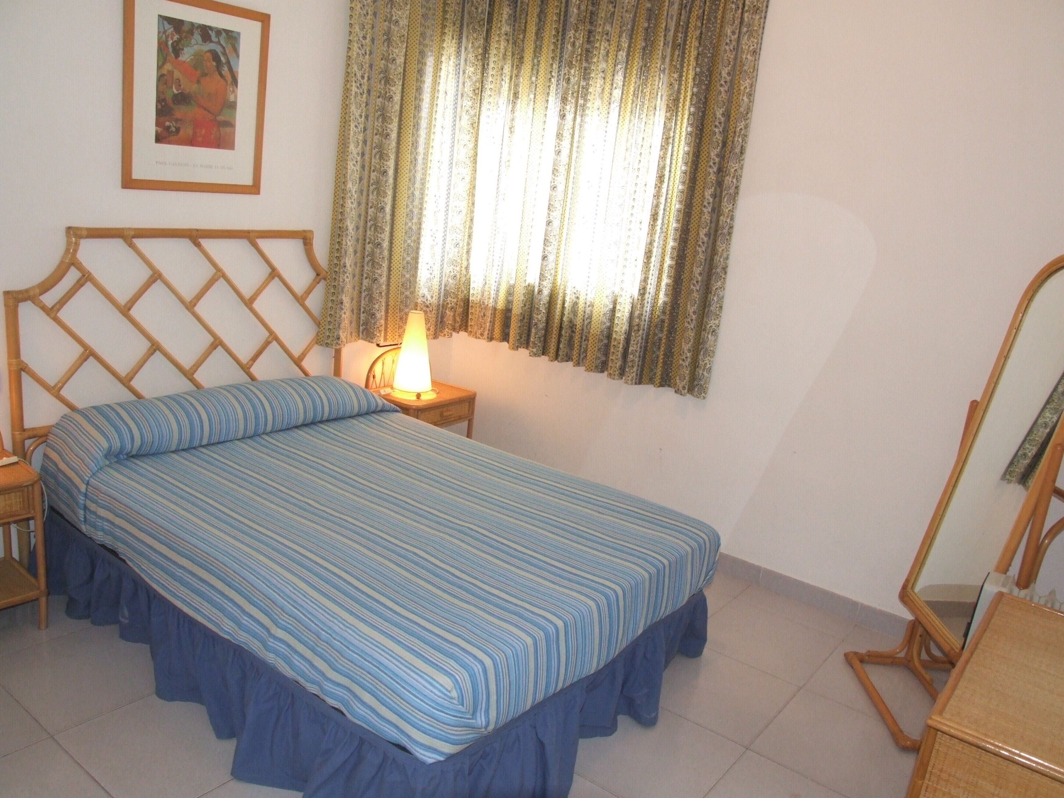 Un dormitorio con cama, mesita de noche, lámpara, espejo y un cuadro en la pared.
