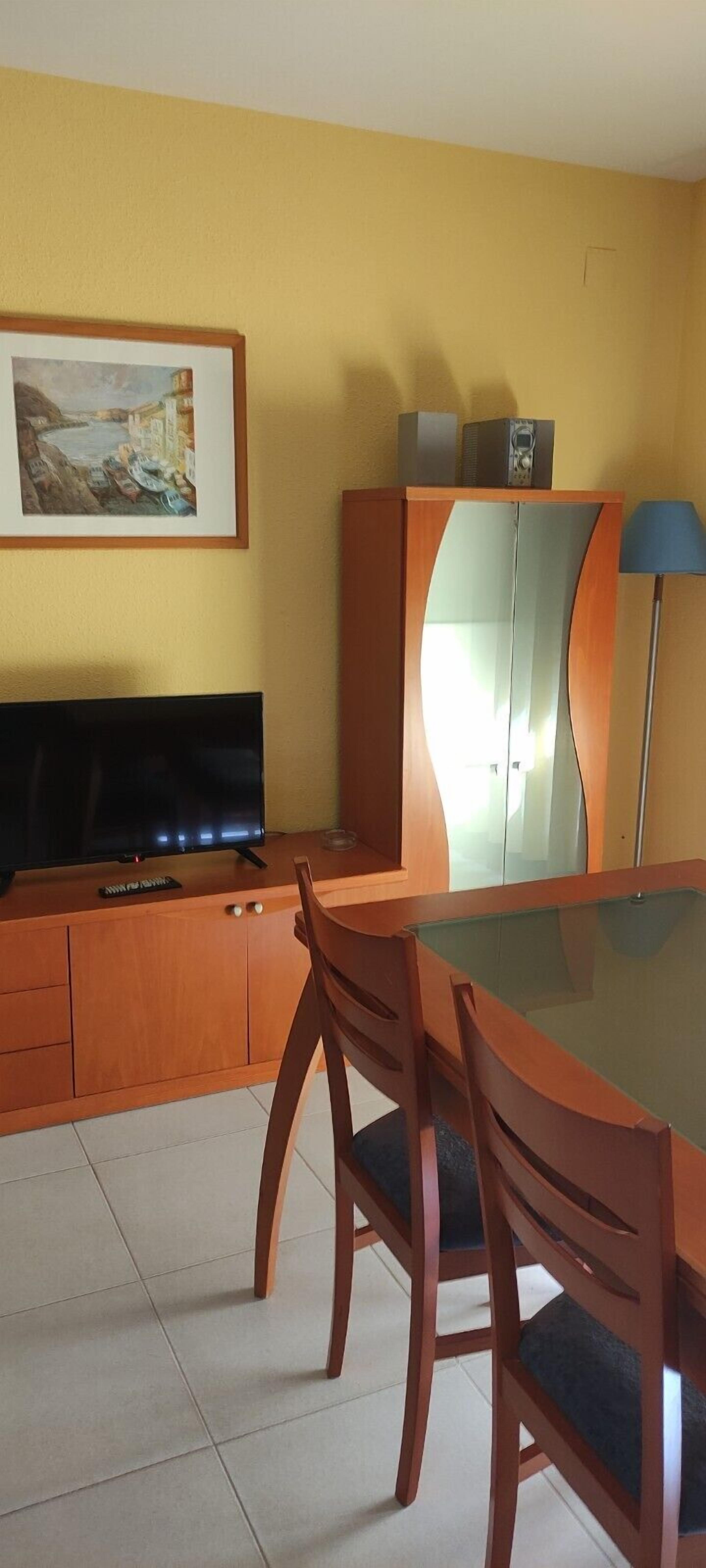 A dining area with a glass table, two wooden chairs, a television on a wooden cabinet, and a framed picture on the wall.