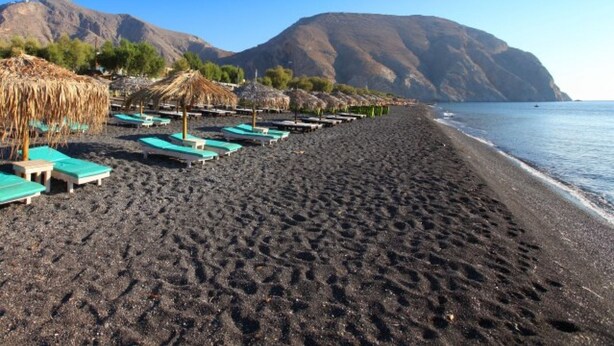 A beach with black sand, rows of blue lounge chairs, and thatched umbrellas. - Perissa Hotel (Santorini)